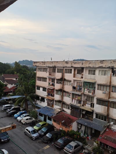 The image shows a high-angle view of a residential area in Ampang Jaya Municipal Council, Malaysia. In the foreground, a concrete overhang is visible on the left. Dominating the scene is a multi-story apartment building with a weathered facade. The building has numerous windows, balconies with laundry hanging out, and metal security bars. Adjacent to the apartment building are smaller, single-story houses with red tile roofs. A parking lot is filled with various cars, including sedans, SUVs, and smaller vehicles. Greenery, including trees and palm trees, is interspersed throughout the scene. The sky is overcast with some hints of sunlight breaking through. The overall impression is that of a densely populated urban residential environment.