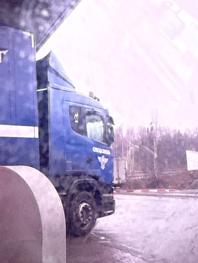 The image displays a large blue heavy-duty truck, possibly a specialized vehicle or a semi-trailer cab, positioned diagonally across the frame. On the side of its cabin, the Cyrillic text "СПЕЦСВЯЗЬ" (SPETSSVYAZ), meaning 'Special Communications,' is clearly visible, accompanied by a stylized eagle or bird emblem. The truck appears to be parked or moving slowly on a paved road or lot.
In the background, a sparse line of trees suggests a rural or semi-urban environment. To the right, a white and red striped barrier post is partially visible. The ground surface exhibits a textured appearance, which could indicate a thin layer of snow, ice, or simply worn asphalt. The overall lighting is very bright and somewhat washed out, casting a high-key, slightly purplish-pink hue over the scene. This bright, diffuse light might suggest an overcast day or highly reflective snow cover.
No people are visible in the scene. A portion of another vehicle or trailer, also in shades of blue and gray with a white stripe, is seen in the lower left and bottom center of the frame, indicating the blue truck might be attached to it or parked adjacently. The setting is Kiselevka, Russia.