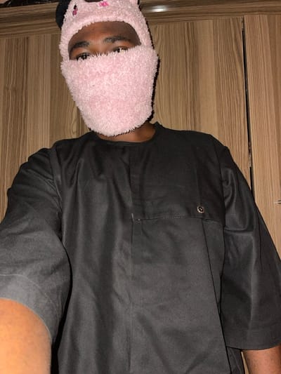 A single individual is depicted in a close-up, chest-up shot, indoors in Unguwar Gabas, Nigeria. The person, presenting a dark skin tone, wears a black, loose-fitting, long-sleeved garment with a rounded neckline. The garment features a distinct seam across the upper chest, incorporating a small, rectangular pocket-like detail on the left and a circular metallic button or snap on the right side. The individual's head is covered by a pink, fuzzy balaclava-style mask that conceals the nose, mouth, and chin, extending over the forehead. Two small, rounded protrusions resembling animal ears are visible on top of the mask. Only the individual's eyes are visible through the mask's opening. A portion of the person's left arm is visible in the lower-left foreground, suggesting the image is a self-portrait. The background consists of vertically oriented, light brown wooden paneling, consistent with an interior wall or cabinetry. The lighting is even but appears somewhat subdued.