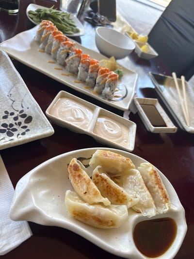 The scene depicts an indoor dining table within an Asian restaurant, consistent with establishments found in Naples, United States. The dark brown table surface holds multiple ceramic dishes. In the foreground, a white, irregularly shaped plate contains five pan-fried dumplings (gyoza), with a separate compartment holding a dark brown dipping sauce. Centrally, a long, rectangular white plate presents ten pieces of a sushi roll, each topped with reddish-orange roe and a light-colored sauce drizzle. Adjacent to the sushi, a two-compartment rectangular dish holds two servings of a creamy, pale sauce. In the background, a white bowl filled with green edamame pods, an empty white bowl, and a small rectangular dish containing lemon wedges are visible. A separate place setting includes a rectangular dish with a dark liquid (likely soy sauce) and a pair of wooden chopsticks. No individuals are present in the frame. The environment suggests natural lighting originating from an unseen window on the right.