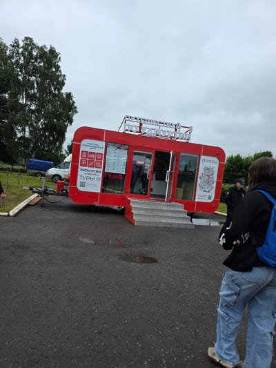 The image shows a red tourist information center trailer labeled "ПУТЕШЕСТВУЙ ПО УДМУРТИИ" (Travel around Udmurtia) situated in the village of Starye Bygi, Russia. The trailer is parked on an asphalt surface, possibly a roadside or parking area, with puddles of water present. A white van and a blue-covered trailer are visible in the background, along with trees and a cloudy sky.
In the foreground, a person wearing a black hoodie, blue cargo jeans, and a blue backpack is facing away from the camera, towards the tourist information center. To the right of the trailer, a police officer in uniform is standing and looking in the direction of the tourist center. Another person's hand is visible on the left side of the image, near a fence. The scene appears to be an outdoor setting, possibly a tourist stop or a public gathering.