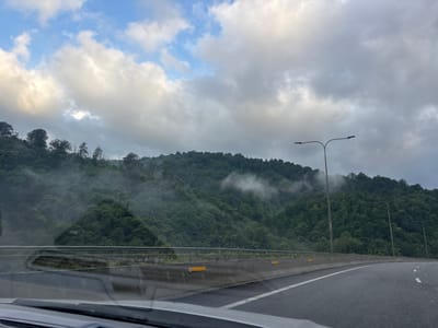 The image depicts a highway curving through a mountainous, forested area, with some fog or mist present on the slopes. The sky is partly cloudy. On the right side of the highway, there are two streetlights. The highway barrier is visible in the foreground, with yellow reflectors. The scene appears to be taken from a moving vehicle. Based on available information, this location is consistent with the highway route near Tsakva, Georgia, which is known for its coastal mountains and lush vegetation. No people are visible in the image.