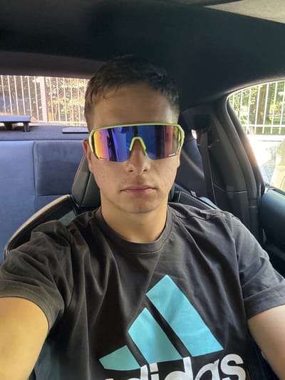 A young man with short brown hair is sitting in the driver's seat of a car, wearing large, neon-green framed sunglasses with mirrored lenses and a dark grey t-shirt with a light blue Adidas logo. The interior of the car is visible, including dark grey seats with red stitching, a seatbelt, and the car's roof lining. Through the rear window, a white metal fence and trees with sunlight filtering through are visible, suggesting an outdoor setting in a city. Given the presence of the Adidas logo, which is a globally recognized sportswear brand, the scene likely takes place in a developed urban area. The car's interior, coupled with the view through the window, indicates the man is parked. The specific location in Cetinje, Montenegro cannot be determined from the visual information alone.