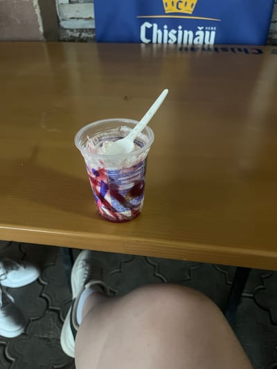 The image shows a close-up of an ice cream cup with a spoon in it, resting on a wooden table. The cup is made of clear plastic and contains a swirl of white ice cream and red fruit topping. Some text is visible on the wrapper of the ice cream cup, but it is mostly obscured. To the left of the table, a blue banner with yellow lettering is partially visible. The banner appears to have text that reads "Chisinau" and "HERE". The overall setting seems to be outdoors, possibly a cafe or a public seating area, given the patterned paving stones visible in the background. A person's legs and feet are visible on the right side of the image, wearing white sneakers and socks. The lighting suggests it could be daytime, but the specific time of day or weather conditions are not discernible from the image. The image is oriented sideways.