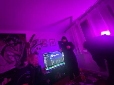 A dimly lit room bathed in purple light features three people. One person sits in front of a large computer screen displaying a music app. Another person, wearing a hoodie, stands in the center. A third person is partially visible on the right side. A large painting and a potted plant adorn the wall. A table in the foreground holds various items, including a bottle. The room's lighting makes it challenging to discern details.