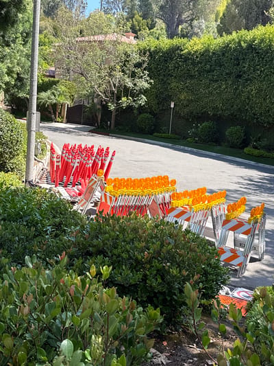This media file depicts a row of orange traffic cones and barricades lined up on a paved surface, likely a residential street in Beverly Hills, California. The cones are stacked in several rows, with the front rows being predominantly orange and the back rows transitioning to yellow tops. Some of the barricades have orange and white reflective stripes. In the background, lush green hedges and trees line the street, suggesting a well-maintained, upscale neighborhood. A portion of a house with a tiled roof is visible through the foliage on the left side of the frame. The lighting suggests it is daytime, with sunlight casting shadows on the pavement, indicating a bright and possibly warm day. The scene is static, with no visible activity or people. A white post with a traffic signal head is partially visible on the left edge of the frame. The overall impression is one of preparation for road work or traffic control in a scenic and affluent area.