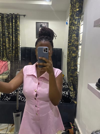 The image displays a female individual taking a mirror selfie inside what appears to be a bedroom. Her face is obscured by a light blue/grey smartphone held at eye level. She possesses dark dreadlocks styled in a high bun. She wears a light pink, sleeveless romper featuring a collar and a front placket with gold-colored buttons.
The room, reflected in the mirror, has light-colored walls. A dark upholstered headboard of a bed is visible behind the individual, with the bed covered by black and white patterned bedding. Above the headboard hangs a framed artwork depicting a stylized grey figure. Dark curtains with abstract gold patterns are drawn across windows on both the left and right sides of the reflection. A metallic pedestal fan stands on the floor to the left of the individual. On the floor in the bottom right, a pair of dark slip-on sandals are visible. A white wall-mounted shelf on the right holds a dark shoe. A small blue object is taped to the wall near the window on the right. This scene is situated within a private residence in Okpanam, Nigeria.