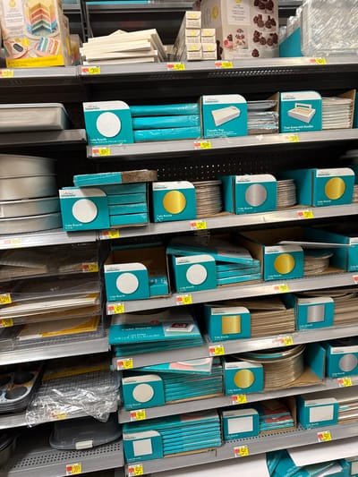 The image displays a well-stocked retail store aisle, specifically a section dedicated to baking and cake decorating supplies. Products are neatly arranged on dark metal shelves, viewed from a slightly elevated perspective.

The primary subjects are various cake boards, treat boxes, and cupcake trays, predominantly from the "Celebrate!" brand. These cake boards are available in multiple shapes (round and square) and finishes (gold, silver, and white), often packaged in distinctive teal or turquoise boxes with cut-out windows revealing the product's color and shape. Stacks of cake boards are visible in different sizes, with many priced at $2.98, $4.97, $5.47, and $6.97, among others.

Other visible items include "Treat Boxes with Handles," priced at $5.98, and "Cupcake Tray" packaging. In the bottom left, various baking pans are stacked, alongside a box for a Wilton brand layer cake set, priced at $15.52. Small packages from the "Wilton" brand are also seen on the left shelf. The overall setting is a clean and organized retail environment, likely a supermarket or craft store in Worcester, United States. There is no specific activity taking place, only products on display for sale.
