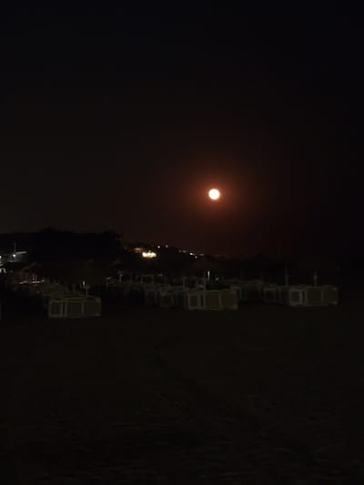 The image captures a nighttime scene in Quarteira, Portugal, with a prominent full moon casting a soft glow. The moon appears reddish-orange, suggesting it's either low in the sky or affected by atmospheric conditions. In the foreground, a Ferris wheel stands silhouetted against the dark sky. The wheel is illuminated with lights, and several enclosed gondolas are visible. Some of these gondolas appear to be white and rectangular. The overall setting is dark, indicative of nighttime. The presence of the Ferris wheel suggests an amusement area or a waterfront attraction, common in coastal towns like Quarteira. There are no visible people or text in the image. The atmosphere seems quiet and serene, dominated by the presence of the moon and the silent structure of the Ferris wheel.