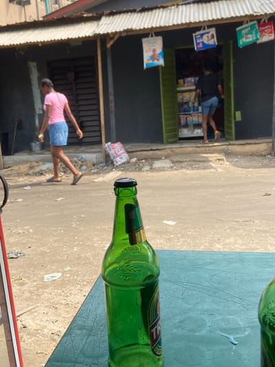 The image depicts a daytime street scene in Alaiyabiagba, Nigeria, viewed from a low perspective. In the immediate foreground, a partially full green glass bottle with a black cap rests on a teal-colored table surface, exhibiting faint liquid spills.
Across an unpaved, debris-strewn road in the midground, two female individuals are observed. One subject, wearing a pink short-sleeved top, a light blue denim skirt, and dark sandals, walks from left to right, carrying a small, light-colored object in her left hand. The second subject, seen from behind in a dark top and light blue shorts, is positioned in the threshold of a small commercial establishment, appearing to enter or exit.
This establishment, situated in the background, features a dark grey facade and open bright green double doors revealing shelves stocked with various products. A corrugated metal roof extends over the entrance, from which multiple advertising bags, including one prominent white bag depicting a baby, are suspended. To the left of this shop, another dark-colored building is visible with a closed, corrugated metal gate. The environment suggests a local commercial area under natural daylight.