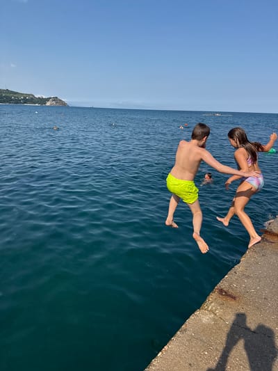 On a bright, sunny day, two children, a boy and a girl, are captured mid-jump as they leap off a concrete pier into the clear blue sea. The boy, wearing bright yellow swim trunks, is to the left and slightly ahead of the girl. The girl, in a multi-colored swimsuit, is just behind him, her body angled as she leaps. Both children are barefoot. In the background, the calm sea stretches out to the horizon under a vast, cloudless blue sky. The coastline of Partenit, Ukraine, is visible in the distance, with green hills and some buildings dotting the landscape. Other people can be seen swimming and enjoying the water further out. A shadow of an adult is cast on the pier, suggesting their presence.