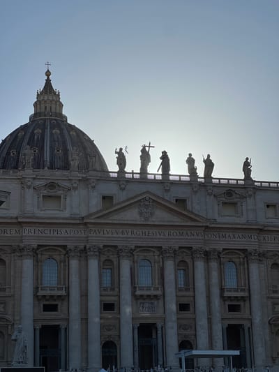 An exterior, backlit, slightly low-angle photograph captures a significant portion of St. Peter's Basilica in Città del Vaticano, Vatican City, under a clear, bright sky. The prominent, ribbed dome, topped with a lantern and cross, occupies the upper left quadrant, appearing partially silhouetted. Below and to the right of the dome, the upper section of the basilica's facade is visible, featuring a balustrade adorned with several large, heavily silhouetted statues of saints and apostles. One statue on the far left clearly holds a cross. Another appears to carry a staff or cross over its shoulder. The sun's position behind the structure creates strong backlighting, causing the figures and the upper dome to appear as dark silhouettes against the bright sky, which transitions from light blue at the top to near white where the sun is obscured. Below the statues, a frieze bears a partial Latin inscription in capital letters: "OREM PRINCIPIS APOST PAVLVS V BVRGHESIVS ROMANVS PONT MAX AN MDCXII," referencing Pope Paul V and the year 1612. The facade below the inscription displays architectural details including large columns, arched windows, and recessed niches, all rendered in muted tones due to the backlighting. At the bottom of the image, indistinct figures of people are visible, gathered behind barriers, and a partial view of a white temporary canopy or structure is present on the right. A large statue of a robed figure, likely St. Peter, is partially visible at the far bottom left.