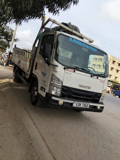 The image features a white Isuzu ELF flatbed truck parked on the unpaved shoulder of a road, captured from a low, front-right angle. The truck is the primary subject, occupying the majority of the frame.
The truck is light-colored, predominantly white, with a dark grille and bumper. The word "ISUZU" is prominently displayed in silver lettering on the front grille. Below the windshield on the passenger side, the model name "ELF" is visible. On the top center of the windshield, blue text reads "M.KORBANI-U/WARE". A small red sticker with "SOYVASA" is located on the upper right side of the windshield. The truck is equipped with a sturdy roof rack, on which some bundled items, possibly tarpaulins or covers, are secured by a wooden frame. The front bumper has two circular yellow auxiliary lights. The license plate is clearly visible on the front bumper, displaying "KDM 760X". The truck's body is a flatbed design with metal side rails, suggesting it's used for transporting goods. On the driver's side door panel, there are several lines of smaller blue text, possibly company information or contact details, but they are too small to be legible.
The setting appears to be an urban or semi-urban area. The truck is parked on a dirt or gravel shoulder, adjacent to a paved road that extends into the background. To the left, a large tree with dense green foliage partially overhangs the scene, providing shade. Behind the tree, a utility pole and faint figures of people are discernible. In the background to the right, a multi-story building with numerous rectangular windows or openings is visible. Further down the paved road, a small blue three-wheeled vehicle, possibly a tuk-tuk, can be seen.
The time of day appears to be daytime, with a bright sky showing some scattered clouds, indicating good visibility. The lighting is even, without harsh shadows, suggesting either late morning or early afternoon. The overall impression is a roadside scene in a populated area. The photo is taken with a slight tilt, making the truck appear angled.