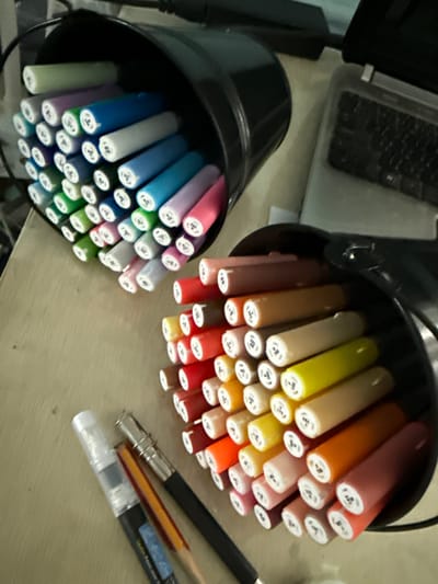The image shows a close-up, overhead view of several markers in black cylindrical holders, arranged on a light-colored, textured surface that appears to be a desk or table. One holder is in the foreground on the left, filled with markers in various shades of green, blue, purple, and white. The markers are angled upwards and outwards, with their white tips visible.
To the right and slightly behind the first holder is another black cylindrical holder, also filled with markers. These markers are in warmer tones, including shades of orange, red, pink, brown, and yellow. They are also angled upwards and outwards, with their white tips showing. Some of the white tips have small black numbers or symbols printed on them.
In the lower right quadrant of the image, there are several writing instruments lying on the surface. One is a black marker with a silver metal ferrule, a sharpened pencil with an orange body, and a clear plastic barrel with a black cap, which appears to be a water brush pen.
The lighting suggests an indoor setting, possibly during the daytime. There are no people visible in the image. The overall impression is one of an art or craft supplies being organized or prepared for use. The location context of Vladivostok, Russia, does not provide any specific visual cues within the image itself.