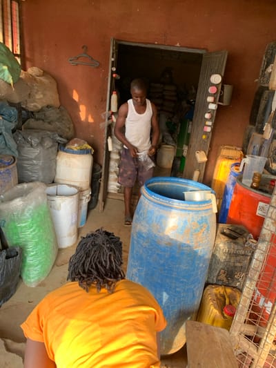 This image depicts an indoor scene in Ube, Nigeria, characterized by a utilitarian and cluttered environment. The reddish-brown walls suggest a non-residential structure, possibly a storage area or a workshop.
In the mid-ground, a male individual stands facing forward, positioned within a doorway that leads to a darker, equally cluttered room. He is wearing a white sleeveless tank top and dark-patterned shorts, with sandals on his feet. His hands are engaged with a clear plastic bag, which he appears to be opening or manipulating. To his left, mounted on the wall, is a dark grey plastic clothes hanger. Along the right side of the doorway frame, a string or wire holds several small, repurposed plastic bottles and containers of various colors, including white, pink, and red.
In the immediate foreground, partially obscured and with their back to the camera, is another individual. This person is wearing a short-sleeved orange shirt and has dark dreadlocks or braided hair. They are bending forward, their exact action not fully discernible.
The scene is dominated by numerous containers. To the left of the doorway, large plastic bags, some opaque grey, some filled with a light-colored substance, are stacked against the wall. A large green translucent bag is also visible on the floor, next to a white plastic barrel. To the right of the central standing figure, a large, cylindrical, weathered blue plastic barrel with an open top stands prominently; a white plastic pitcher or scoop is visible inside its rim. Surrounding this blue barrel are various other containers, including yellow and black jerrycans, a red barrel, and additional stacked tires or dark, cylindrical objects. A clear plastic pitcher with a handle is visible on top of one of the red containers on the right side.
Natural light appears to enter from an unseen source to the upper left, casting distinct shadows on the floor and objects. The overall impression is a busy, functional space dedicated to storage or processing of bulk materials.