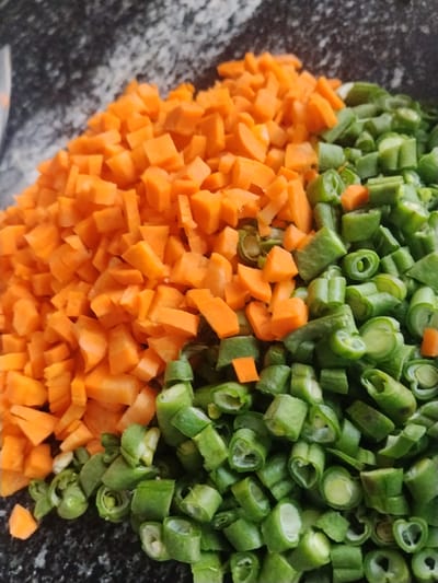 A close-up, top-down view captures a pile of chopped vegetables, predominantly diced carrots and cut green beans, resting on a speckled dark grey surface. The carrots are a vibrant orange, cut into roughly uniform cubes. The green beans are sliced into short, circular pieces, revealing their hollow interior and seeds. The vegetables are sectioned, with the carrots on the left and the green beans on the right. The background appears to be a countertop or table. There are no people or discernible elements that would indicate a specific location within Mangu Town, Nigeria.