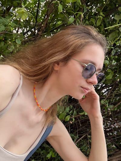 A young woman with light brown hair is resting her chin on her hand, looking down and to the side. She is wearing large, round sunglasses with a light colored frame and a beaded necklace in shades of orange and yellow. The background is filled with lush green foliage and branches, suggesting an outdoor setting. The woman is wearing a light-colored tank top, and part of a blue strap is visible. The scene appears to be taken in Kragujevac, Serbia, potentially in a park or garden area.