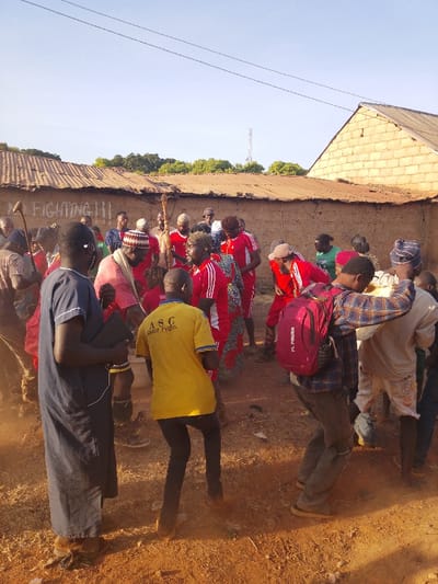 A group of people gather outside a mud brick structure with a corrugated metal roof. Many wear red athletic gear with white stripes on the sleeves. Others are dressed in various casual attire, including a yellow shirt with black lettering, a blue robe, and plaid shirts. Some hold sticks. The ground is dusty, and a few trees are visible in the background. The scene suggests a village setting with people engaged in some form of event or gathering.