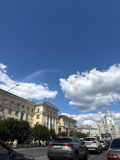 The image depicts a street scene in Voronezh, Russia, under a bright blue sky with scattered cumulus clouds. The perspective is from within a line of cars, suggesting the photo was taken from a vehicle. The cars are stopped, possibly in traffic.

The foreground is dominated by the rear of a dark-colored SUV. Further down the street, other vehicles are visible, including a white SUV with a license plate that reads "H085HP 105". The buildings lining the street are multi-story, with a pale yellow facade on the closer building, featuring numerous windows and arched detailing. Across the street, a large, ornate white building with spires stands prominently against the sky, indicative of historical architecture. Trees with dense green foliage line parts of the sidewalk.

Several people are visible on the sidewalk, some appearing to be waiting or walking. One group of people is gathered near a fence or barrier. The scene suggests a daytime activity, with the sunlight casting shadows and illuminating the buildings. The weather appears to be pleasant, with a clear sky and clouds. The presence of cars and pedestrians indicates an urban environment and normal daily life. There is no explicit indication of a specific event or emotional tone beyond a typical urban street scene.