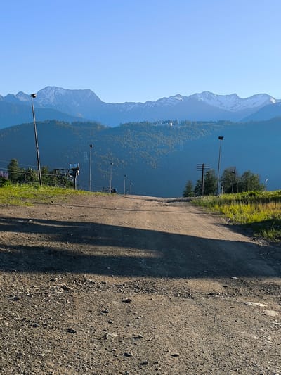 The image depicts a dirt road ascending a grassy slope, leading towards a range of mountains. The road is partially shaded by trees. In the mid-ground, a ski lift and several lamp posts are visible. The mountains in the background are snow-capped and appear majestic against a clear blue sky. The scene is set in Estosadok, Russia, a mountainous resort area. The image captures a serene and picturesque landscape.