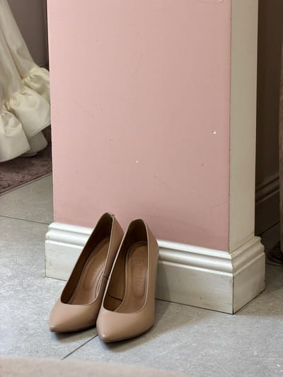 A pair of beige, pointed-toe high heels are placed on a light gray tiled floor. The shoes are positioned near a corner where a pink wall meets a white, decorative baseboard. The heels are slightly overlapping each other, with the front of the lower shoe visible and the back of the upper shoe visible. The interior of both shoes has a label that reads "GEPUR SHOES". The lighting suggests it is daytime, and the indoor setting appears to be a room with a floor and wall. The overall scene is simple and static, with no people or active events depicted.
