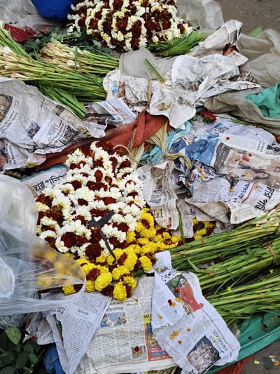The scene depicts a ground-level area, likely a flower stall or vendor's display, in Bardhaman, India. No individuals are present. The surface is covered with crumpled Bengali newspapers, acting as a base for various floral items.
Prominently featured are multiple garlands. At the top-center, a large, rounded garland composed of white and dark red/maroon flowers is visible. A similar garland rests in the mid-left. In the bottom-center, a pile of bright yellow marigold garlands and loose marigold blossoms is present. Resting on the white and maroon garland in the bottom-center is a pair of dark-colored scissors with a distinct curved handle.
Bundles of long, green flower stems, possibly tuberoses or gladiolus, are arranged along the left side and extending to the bottom-right. Dispersed among the newspapers and flowers are fragments of reddish mesh fabric and transparent plastic sheeting. A dark blue fabric or tarp is partially visible in the top-left corner. The environment appears utilitarian, consistent with a busy market setting.