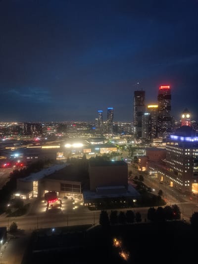 An aerial, nighttime view showcases the Square One City Centre area in Mississauga, Canada. The scene is dominated by a dark blue sky with some faint clouds. The city below is illuminated by the lights of various buildings and streetlights. Prominently featured are several skyscrapers, including two very tall buildings in the center-right, one topped with red lights and another with yellow lights. To the left of these, two other tall buildings display blue lights. In the foreground-left, a large, modern building with a red "PARKING" sign illuminated above its entrance is visible. Further to the right, a building with a clock tower and blue lighting stands out. The image captures the vibrant urban landscape of Mississauga at night, with a clear focus on its modern architecture and city lights.
