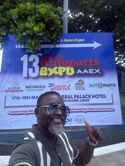 An adult male is prominently featured in a selfie, smiling broadly with visible teeth and giving a "thumbs up" gesture with his left hand. He is dark-skinned, has a graying beard, and wears reflective aviator-style sunglasses and a short-sleeved collared shirt with olive green and white vertical stripes. Behind him is a large blue and white banner advertising the "13th Africa Autoparts EXPO AAEX" and the "20th Lagos Motor Fair." The banner specifies the event dates as "17TH-19TH MAJ" and the venue as "FEDERAL PALACE HOTEL VICTORIA ISLAND, LAGOS," along with various organizational logos, including ALCMAN, MOMAN, and AUTOPARTS Nigeria. Green foliage from a tree is visible above and to the left of the banner, partially obscuring its top. A light pole stands behind the tree. The general location for this scene is Itirin, Nigeria.