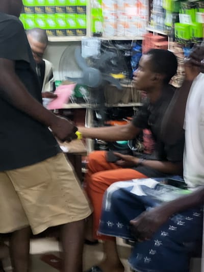 In a shop at Idimu, Nigeria, two individuals are present. One individual wearing an orange garment and a blue garment with white patterns is standing behind another. The other person is wearing a black garment and tan shorts, and a yellow object is in their hand. An oscillating fan is visible in the background along with shelving filled with assorted items.