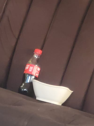 A Coca-Cola bottle lies on a dark brown surface next to a white bowl. The location, Rafin Bauna, Nigeria, is not inferable from the image.