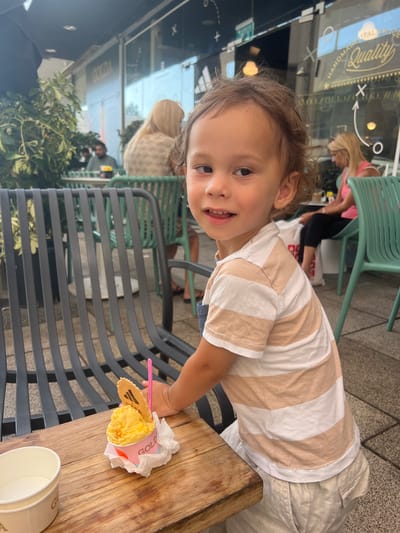 A young child with curly brown hair and wide eyes smiles at the camera, their mouth open as if speaking. They are wearing a striped t-shirt with wide horizontal bands of beige and white. The child's right hand, small and chubby, reaches towards a cup of bright yellow ice cream. A pink plastic spoon is inserted into the ice cream, and a waffle cookie with a patterned design is perched on the edge of the cup. The ice cream is in a pink paper cup with white accents and appears to have the word "GOLD" visible in white lettering.
The child is leaning over a weathered wooden table. Behind them, a dark metal chair with a slatted design is visible. In the background, blurred figures can be seen in what appears to be a cafe or restaurant setting. Through a glass window, text can be partially read, including "Quality" and "Italian Market" in elegant script, suggesting an Italian food establishment. Green chairs are also visible in the background. The lighting suggests it is daytime, possibly sunny. The child's expression is joyful and engaged. The overall scene is casual and appears to be taking place outdoors or in a semi-outdoor seating area. The location context provided is Kiryat Bialik, Israel.