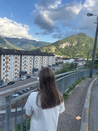 The image, taken from an elevated perspective, shows a person's back as they look out over a town nestled at the base of a lush, green mountain. The person has long, brown hair and is wearing a white t-shirt. In the foreground, a metal railing runs along the edge of what appears to be a walkway or bridge.
Below the walkway, a street lined with parked cars runs alongside a row of apartment buildings. These buildings are characterized by their uniform architecture, with light-colored facades and repeating patterns of windows, some of which have orange frames. The street itself has a visible yellow curb and some cracks in the asphalt. A small orange and white item, possibly litter or a marker, is visible on the road.
The background is dominated by a large, verdant mountain, its slopes covered in dense trees. The sky above is a mix of bright blue and dramatic, greyish-white clouds, suggesting either changing weather or a partly cloudy day. A street lamp is visible attached to a pole in the upper left quadrant of the image, near the mountain.
The overall scene suggests a residential area in a mountainous region. The lighting indicates it is likely daytime, possibly late afternoon given the quality of light. The person's posture and the vastness of the landscape suggest a moment of quiet observation or contemplation. The provided location context places this scene in Saint-Claude, France. No discernible text is visible in the image.