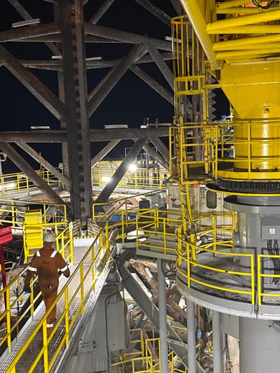 The image shows an industrial scene at night, possibly an oil rig or a similar offshore platform, with a person in an orange jumpsuit walking on a metal walkway. The structure is characterized by large, dark metal beams and bright yellow railings and ladders. In the background, there are yellow industrial structures and equipment, some of which appear to be machinery. The lighting suggests artificial illumination from within the structure. There is no visible indication of the city of Unknown, Italy.
