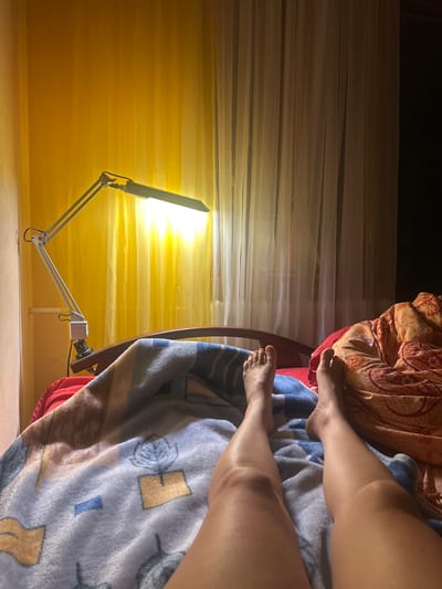 The image shows a person's legs and feet in the foreground, lying in a bed. They are covered with a blue and white patterned blanket on the left and a rust-colored blanket with a paisley pattern on the right. The scene is dimly lit, with a bright yellow curtain in the background illuminated by a long, adjustable desk lamp positioned on the edge of the bed. A dark curtain or wall is visible to the right. The setting appears to be a bedroom.