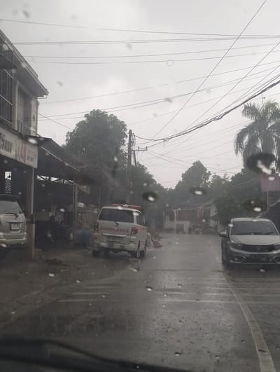 The image depicts a street scene in Banda Aceh, Indonesia, during a heavy downpour. The weather is overcast and rainy, with numerous water droplets visible on the foreground, suggesting the photo was taken from inside a vehicle.
In the foreground, the road is wet and reflects the dim light. To the left, part of a white SUV is visible. Further down the street, an ambulance with red and white markings and flashing lights is parked. The word "AMBULANCE" is clearly visible on its side. A license plate reads "BL 9234 LB". Next to the ambulance, there are some people sitting under a covered structure.
On the right side of the street, a silver car is parked. The street is lined with buildings and trees, which appear lush and green, indicating a tropical environment. Power lines are strung across the sky, a common sight in urban and semi-urban areas. The overall atmosphere is somber due to the rain and the cloudy sky. The presence of the ambulance suggests a possible emergency or a medical transport situation. The time of day is unclear due to the overcast conditions, but it appears to be daytime.