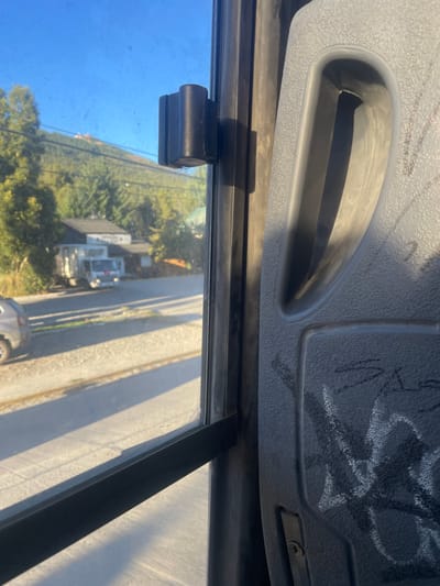 The image captures an exterior view from inside a moving or parked vehicle, likely a bus, through a window on a clear, sunny day in San Carlos de Bariloche, Argentina.

**Interior Details:**
To the right, a portion of a grey, textured plastic seat back is visible, featuring an integrated handle-like recess and white, graffiti-style markings. A black vertical window frame runs along the right side of the window, with a black, ribbed cylindrical knob affixed to it at eye level. A horizontal dark bar crosses the lower section of the window. The window glass appears lightly smudged.

**Exterior Details:**
Outside, under a clear blue sky, a paved road with distinct long shadows indicating direct sunlight extends across the mid-ground. On the left side of the road, the rear section of a light-colored SUV is partially visible. Further down the road, a white box truck with red accents is parked or stopped in front of a two-story building with a light-colored facade and a dark roof. The background features a verdant, tree-covered hillside ascending towards the horizon. Structures are discernible on the upper slope of the distant hill, consistent with mountain infrastructure. Overhead power lines are visible against the sky and trees in the upper left. The general environment of forested mountains and residential/commercial structures is characteristic of San Carlos de Bariloche, Argentina.