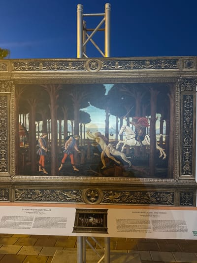 The image displays a large, ornate framed replica of a Renaissance painting set against a twilight sky. The painting depicts a scene in a dense forest with several figures. In the foreground, two young men in traditional attire, one with his head bowed and the other looking down at a stick, walk amongst tall trees. To the right, a pale, nude female figure is being pulled by a white dog, with a distressed expression and reaching arms. Behind her, a white horse with a rider in a red cloak gallops forward, the rider holding a sword aloft. In the background, a fortified structure with tents is visible on the left, and a body of water with ships and hills forms the horizon. The replica is positioned outdoors, with metal scaffolding visible behind and to the sides of the artwork. A descriptive plaque below the artwork, partly obscured by a metal support, identifies the painting as "The Story of Nastagio degli Onesti" by Sandro Botticelli and Studio, and mentions its location in the city of Mazarrón, Spain. The plaque also contains text in Italian and English describing the artwork and its narrative.