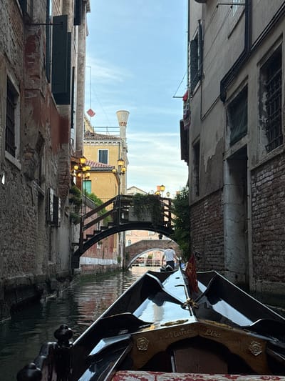 The image depicts a forward-facing view from inside a gondola navigating a narrow canal in Venice, Italy. The gondola's ornate bow, featuring a reflective black surface and gold accents, is prominent in the lower foreground, with the name "Mona Lisa" inscribed on its decorative wooden section. A small red flag with a golden emblem is affixed to the bow.
The canal water is dark green, reflecting the surrounding architecture with visible ripples. Tall, aged buildings constructed of brick and plaster line both sides of the canal. The building on the left exhibits exposed red brickwork and weathered plaster, with dark closed shutters on windows. The building on the right is predominantly light-colored plaster, also showing signs of age, with grates on some windows and clothes hanging on a line high up.
In the mid-ground, a pedestrian bridge with an arched metal structure and wooden steps spans the canal. Ornate, warm-toned streetlights are illuminated on this bridge and on a yellow building beyond it. Green foliage and planters adorn the bridge. A person wearing a white t-shirt and a light-colored hat is visible walking under this bridge, facing away from the viewer. Further down the canal, another stone arch bridge is visible. The sky above is a light blue with some scattered clouds. Typical Venetian chimneys and an antenna are visible on distant rooftops.