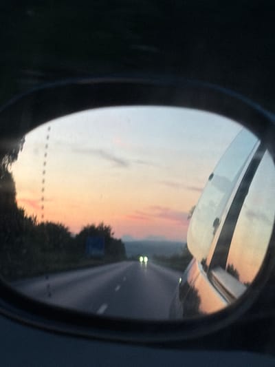 The image is a photograph taken from inside a vehicle, reflected in a side-view mirror. The setting appears to be a highway in Bulgaria, possibly near the town of Ignatievo, during sunset or twilight. The mirror reflects the road ahead, the sky, and the surrounding landscape. Two sets of headlights from an oncoming vehicle are visible in the distance on the road. The sky displays a gradient of colors, with hues of pink, orange, and blue. Trees and foliage line the sides of the road. The reflection in the mirror also shows the vehicle's side and part of the body, with the sunset colors reflecting on its surface. The overall mood is serene and evocative of travel.