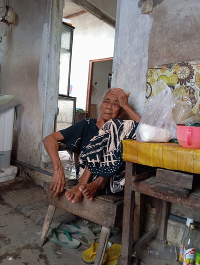 An elderly woman is sitting on a low wooden structure in what appears to be a humble dwelling in Baron, Indonesia. Her feet are bare, and she is touching her forehead with her hand. Near the woman's head are plastic bags filled with what appear to be rice or flour, and a box wrapped in yellow tape is positioned nearby. The surrounding environment includes a worn concrete floor, a stack of flip-flops, and a visible window frame. The interior walls are weathered and simple. A patterned fabric hangs on the wall behind the woman.