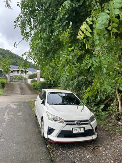 A white Toyota Yaris is parked on the side of a wet, asphalt road in Kathu, Thailand. The car is facing towards a large, white house with a dark tiled roof, accessible via a concrete driveway with an ornate metal gate. The scene is heavily shaded by lush green trees and foliage, with a hilly, vegetated landscape visible in the background. The sky is overcast. The car's license plate reads "กข 7512".