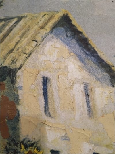 This image is a close-up photograph of a painting, rendered in an impressionistic or textured style. The painting primarily depicts the upper section of a building, likely a house, set against a light background.
**Subject:** A gabled roof, colored in shades of light yellow and tan with darker linear accents, dominates the top portion. Beneath it, the building's facade is depicted in a light beige or cream color. This wall exhibits significant texture and numerous areas of paint flaking and cracking, exposing the underlying canvas weave. Two dark blue vertical rectangular shapes are painted on the facade, suggestive of windows or shadowed openings. The left side of the building features darker, amorphous shapes in tones of brown and deep green, which could represent foliage or shadow. The background in the upper right quadrant is a light blue-grey, consistent with a sky.
**Objects:** The primary object is the depicted building within the painting. The photograph captures the physical texture of the paint and the canvas.
**Actions/Interactions:** No people or explicit actions or interactions are visible in the scene.
**Environment:** The depicted environment is a generalized outdoor setting with a building and a sky. The painting itself does not contain specific geographical markers or unique architectural details that would allow for the precise identification of a specific street, neighborhood, or exact location within the city of Sucina, Spain. The building appears as a generic representation within the artwork.