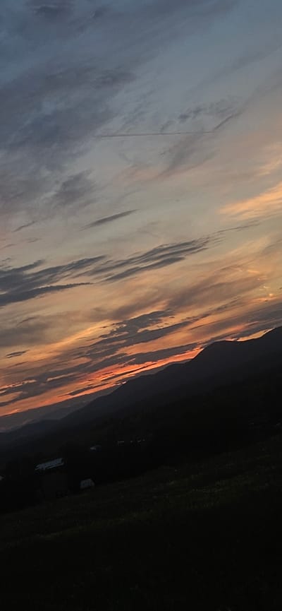 The image depicts a dusky sky over rolling hills, with the sun having recently set. The sky is a gradient of blues, grays, and oranges, with wispy clouds streaking horizontally. In the foreground, dark silhouettes of mountains recede into the distance. The lower portion of the image shows a dark, grassy landscape with faint outlines of buildings or structures, possibly a small village or rural settlement. The scene conveys a sense of natural beauty and tranquility, characteristic of a rural or mountainous region at dusk. The specific location in Momin Prohod, Bulgaria, is not discernible from the visual elements of the image alone.