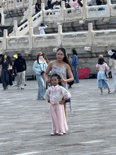 In Dongcheng District, China, at an outdoor location with marble architecture, a woman in a patterned dress stands with a young girl in a traditional pink outfit. The woman has her hand on the girl's back, while the girl stands with her hands on her hips. Several other individuals are visible in the background, some walking, some standing, and one taking a photo. A red umbrella lies on the ground near a person wearing a light blue jacket and jeans, who is holding a camera. The ground is paved with wooden planks, and the architectural elements suggest a historical or monumental site.