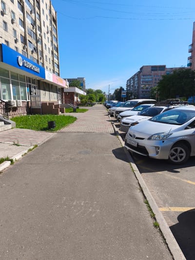 The image captures a street scene in Komsomolsk-on-Amur, Russia, on a clear, sunny day. The perspective is at street level, looking down a sidewalk with parked cars on the right. On the left, a multi-story apartment building with numerous windows and air conditioning units is visible. A bright blue sign with white Cyrillic lettering, "ГАЗПРОМБАНК" (Gazprombank), is prominently displayed on the building's facade, indicating a bank branch. Shops with large windows are situated at the base of the building.
The sidewalk is paved with interlocking bricks. A grassy area with a dark cylindrical trash receptacle is located between the sidewalk and the building. Beyond the sidewalk, several cars are parked in a row along the street. The closest car is a silver Toyota Prius, with its license plate visible as "Р242АХ 27" (R242AX 27). Other vehicles, including a white van and several sedans, are also parked.
Across the street, more apartment buildings and trees are visible, contributing to the urban environment. Utility lines and poles are present in the background. The sky is a clear, bright blue, with a few faint lines that might be wires or power lines. The overall impression is of a typical day in a residential and commercial area of the city. There are no people visible in the image.