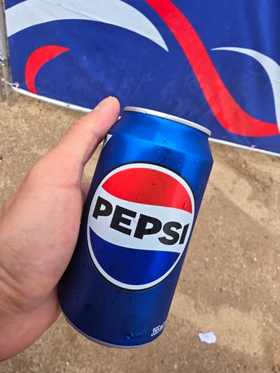 A person's hand is holding a cold, condensation-covered blue Pepsi can. The can has the iconic Pepsi logo, featuring red, white, and blue circles, with the word "PEPSI" prominently displayed in white letters. In the background, a blue banner with red and white wave-like designs is visible. The ground appears to be sandy or dirt-like. The scene is outdoors, likely in an urban environment given the presence of a banner, suggesting a public event or advertisement. There is no specific indication of the location within Daejeon, South Korea, beyond it being an outdoor space.