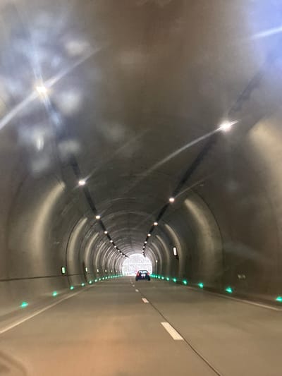The image depicts the interior of a tunnel, likely a modern subway or transit tunnel, with a perspective looking down the length of it towards a bright opening. The tunnel walls are smooth and appear to be made of concrete or a similar material. Along the left and right sides of the tunnel, there are rows of lights. The lights on the left are brighter, reflecting off the curved walls, and appear to be white or warm-toned. On the right side, there's a distinct line of small green lights, possibly markers or indicators.
Towards the far end of the tunnel, a vehicle, possibly a train or a service vehicle, is visible, its red taillights or brake lights casting a faint glow. There are also some white rectangular signs mounted on the walls at intervals, though the text on them is illegible. The overall impression is one of functional, contemporary infrastructure. The lighting suggests it could be either daytime or nighttime, as the artificial lights are prominent. There are no people or animals visible. The setting appears to be underground or enclosed, a transit passage designed for movement. The location context is provided as Surami, Georgia.