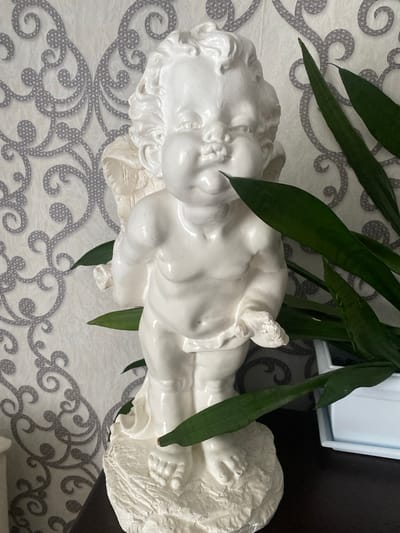 A white ceramic statue of a cherub stands on a textured base. The cherub is depicted with chubby cheeks, curly hair, and delicate wings. It is positioned in front of a wall with a gray ornate pattern. Several green leaves from a plant partially obscure the statue. The background appears to be an indoor setting. There is no identifiable information indicating this location is in Старая Вичуга, Russia.