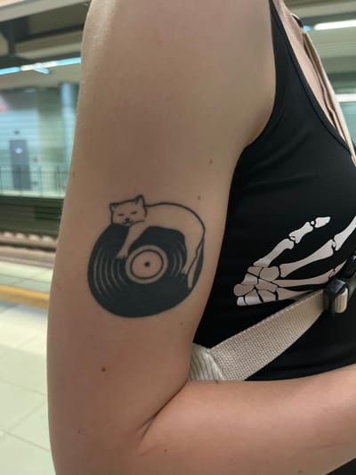 The image shows a close-up of a person's arm with a tattoo of a cat sleeping on a vinyl record. The tattoo is black and located on the upper arm. The person is wearing a black tank top with a white skeleton ribcage design. A light-colored strap is visible across the chest. The background appears to be an indoor public space with horizontal lines and possibly windows or lights. The lighting is bright, and the focus is on the arm and tattoo. The location in Pancharevo, Bulgaria is not discernible from the image.