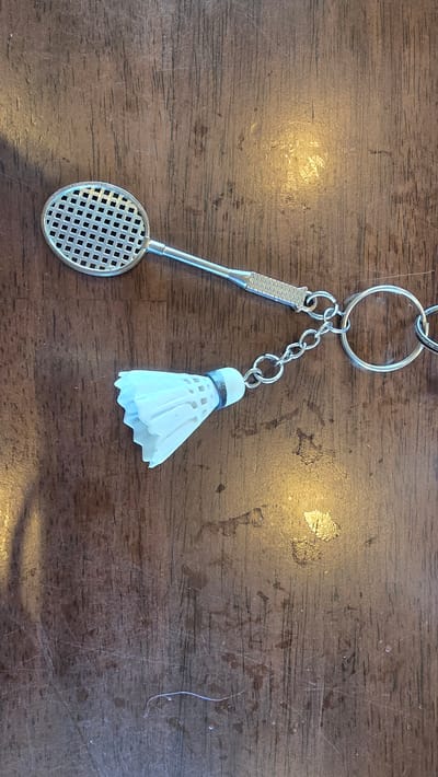 A close-up, top-down shot shows a keychain shaped like a badminton racket and shuttlecock resting on a wooden surface. The racket, made of a silver-toned metal, features a grid pattern on its head and a textured handle. It is connected by a chain to a small, white shuttlecock, also made of metal, with a silver band around its base. The wooden surface appears to be a tabletop with visible grain, scratches, and a few water spots, illuminated by warm, overhead lighting. The background is out of focus, emphasizing the keychain. This image was taken in Choeng Mon, Thailand.